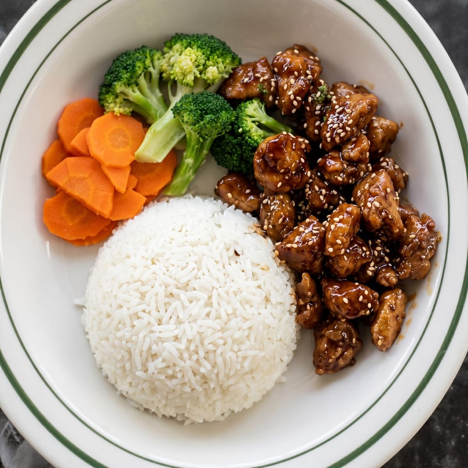 A white bowl with green stripes holds sesame chicken, white rice, steamed broccoli, and sliced carrots.
