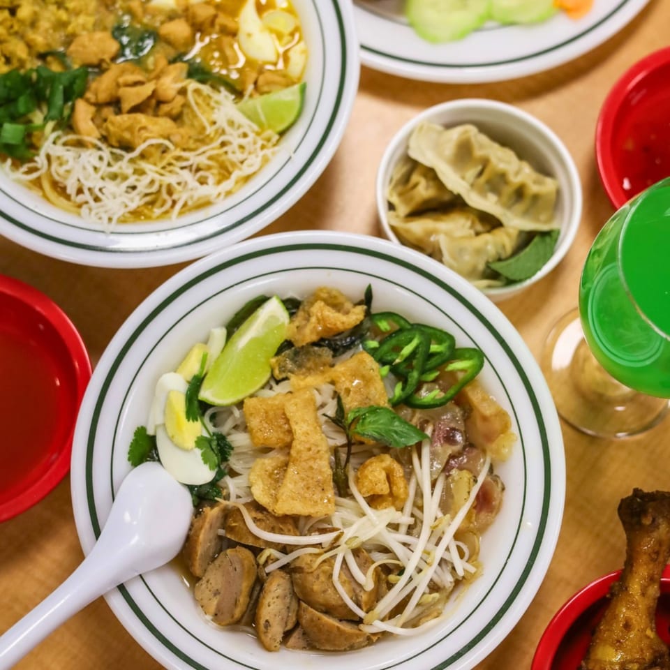 Overhead view of two bowls of noodle soup, dumplings, and a green drink on a wooden table.